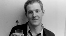 A young white man in his 20s with light hair wearing a dark jacket and holding a cocktail glass.