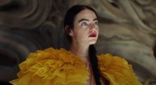 A woman with long, straight dark hair wears red lipstick and a yellow dress with exaggerated, frilly shoulder accents.