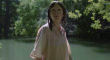 A woman with medium-length, straight black hair wears a light pink blouse and stands in front of a pond surrounded by trees.