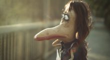 A puppet of a short monster, with a pronounced beak and long brown hair, looks over a bridge and off into the distance.