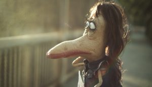 A puppet of a short monster, with a pronounced beak and long brown hair, looks over a bridge and off into the distance.