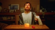 A pre-teen boy sits at a wooden table with one candle lit. He holds a ceramic hand. His face is bloody and his eyes are black.