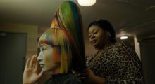 A woman with long, rainbow-colored hair in a tiered high ponytail sits at a salon chair with another woman wearing cheetah print and tightly coiffed hair works on the other woman's hair.