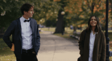 A man dressed in a tuxedo and a woman with a gray coat walk in a park during autumn.