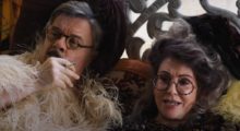 A middle-aged man and woman wear elaborate feathered garb and eccentric glasses recline as the man smokes a cigarette.