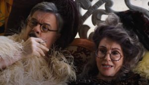 A middle-aged man and woman wear elaborate feathered garb and eccentric glasses recline as the man smokes a cigarette.