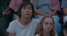 A woman in a white t-shirt sits behind a young girl wearing glasses.