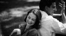 A black and white image of a man and a woman sitting back to back. The woman laughs while the man smiles and smokes a cigarette.