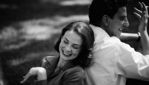 A black and white image of a man and a woman sitting back to back. The woman laughs while the man smiles and smokes a cigarette.