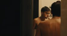A trans man stands in front of a mirror shirtless brushing his teeth after a shower.