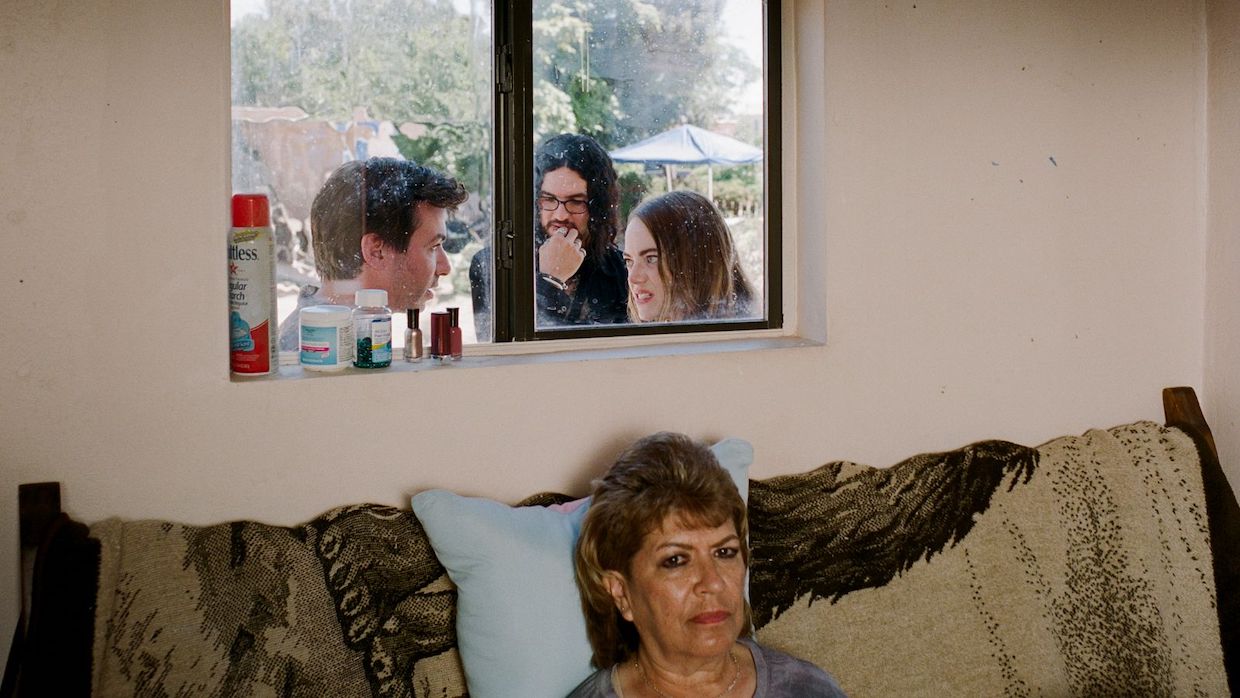 A woman sits on a couch with a blank expression as three people huddle outside, seen through the window right behind her.