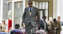 A man in a stylish gray suit walks down the street. He has cropped gray hair and wears black sunglasses.
