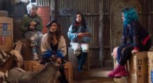 Four women sit in a storage room. They wear brightly colored clothes and accessories, but look mildly bewildered.
