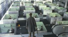 A man wearing a hat and raincoat overlooks rows of symmetrical office cubicles below.