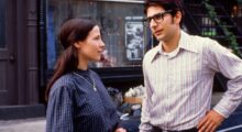 A young woman with long brown hair, blue dress and gold pendant and a taller young man with dark hair, a striped shirt and glasses.