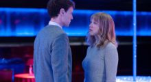 A blonde woman and a man with cropped brown hair stand to face each other as blue lighting illuminates a tank in the background.