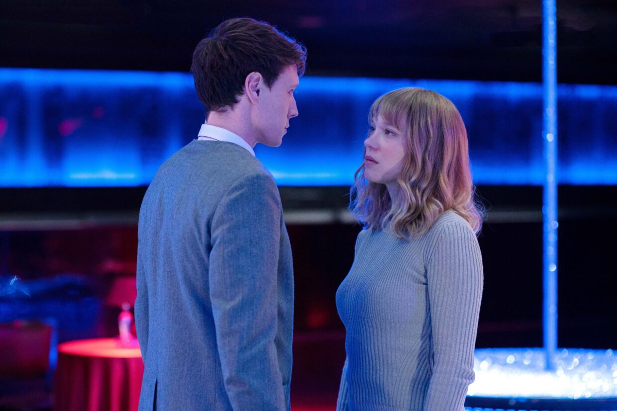A blonde woman and a man with cropped brown hair stand to face each other as blue lighting illuminates a tank in the background.