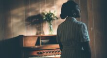 A woman stands in front of an upright piano with a vase of flowers resting on top of it.