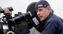 A man in a backwards baseball cap stands next to a 35mm film production camera.
