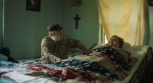 An older man sits at the bedside of a woman lying on a bed, with a crucifix on the wall between them.