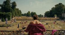 A man in a bathrobe stares out at a large trashed lawn littered with the remains of a party.