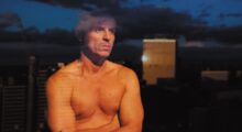 A shirtless man stares out a window at a cityscape