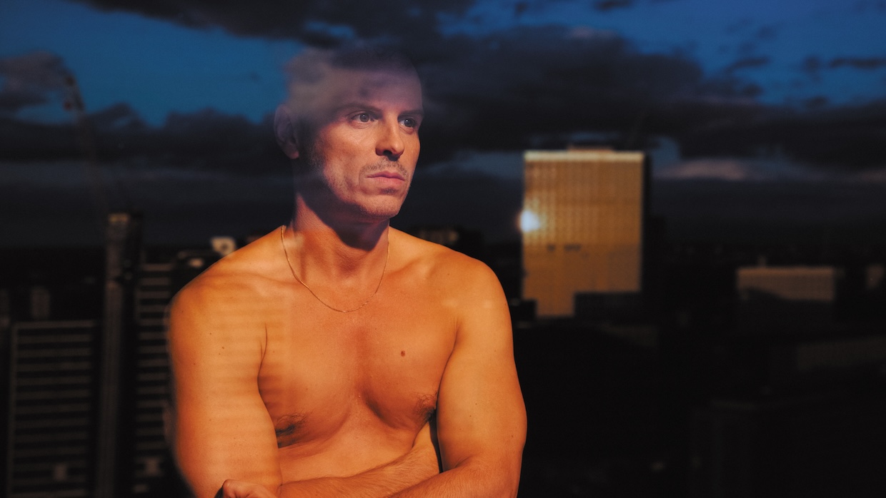 A shirtless man stares out a window at a cityscape