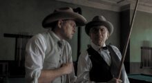 Two white men in cowboy hats stand next to each other. The one on the right holds a rifle.