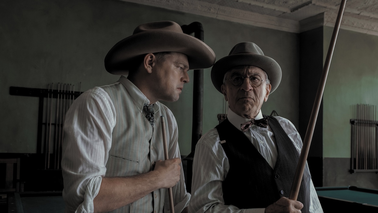Two white men in cowboy hats stand next to each other. The one on the right holds a rifle.