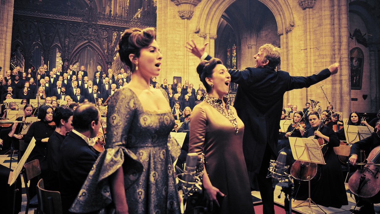 Two women sing as a man conducts a full orchestra behind them in a church.