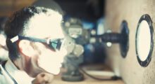 A man in lab glasses stands in front of a porthole, through which a blinding light shines.