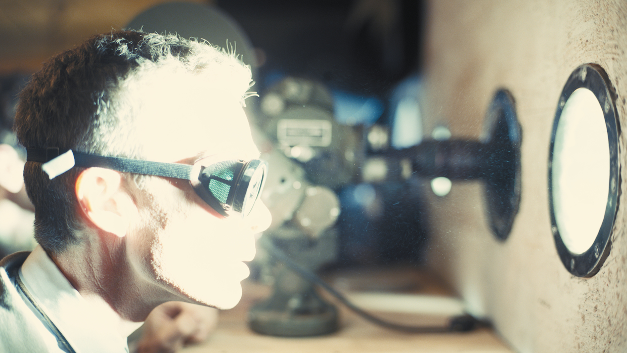 A man in lab glasses stands in front of a porthole, through which a blinding light shines.