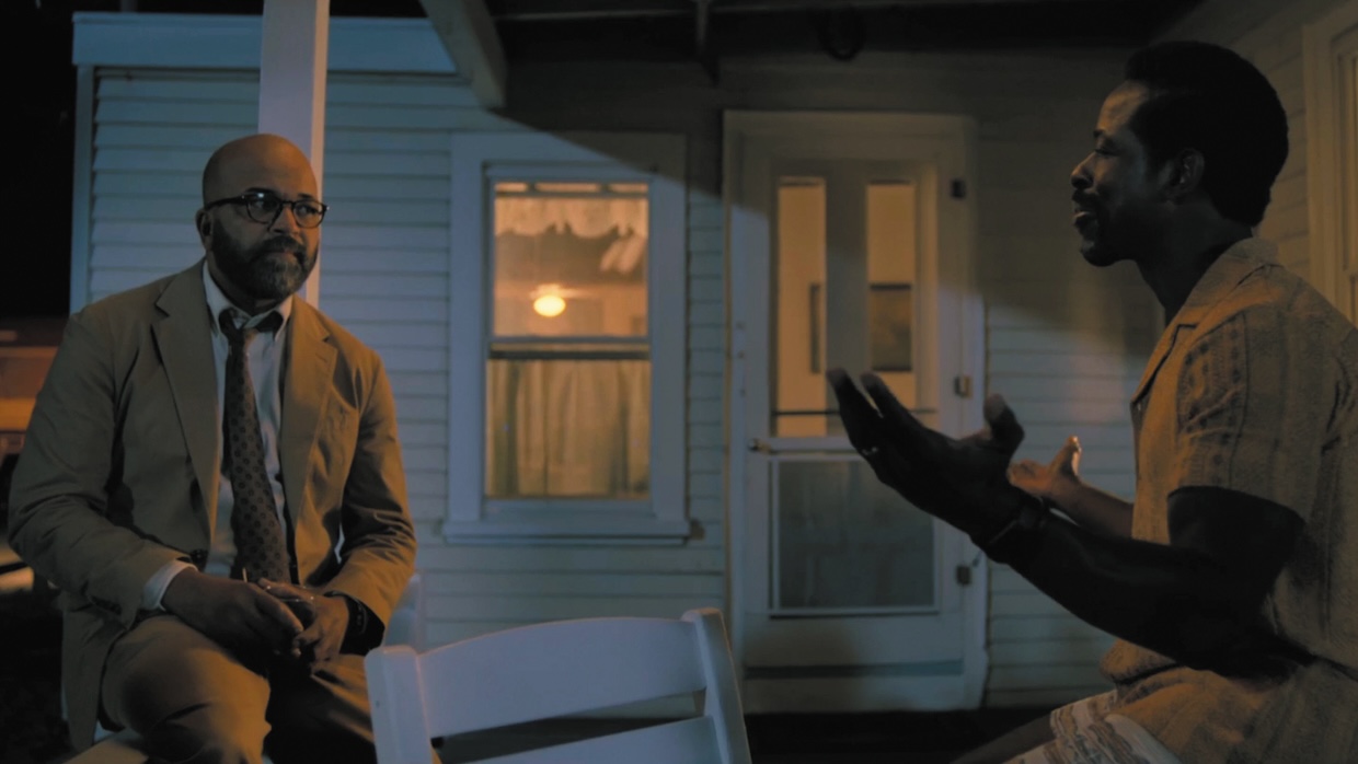 Two black men sit and talk on a porch at night.