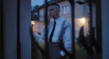 A man in pinstripe suspenders stands outside a house smoking a cigar.