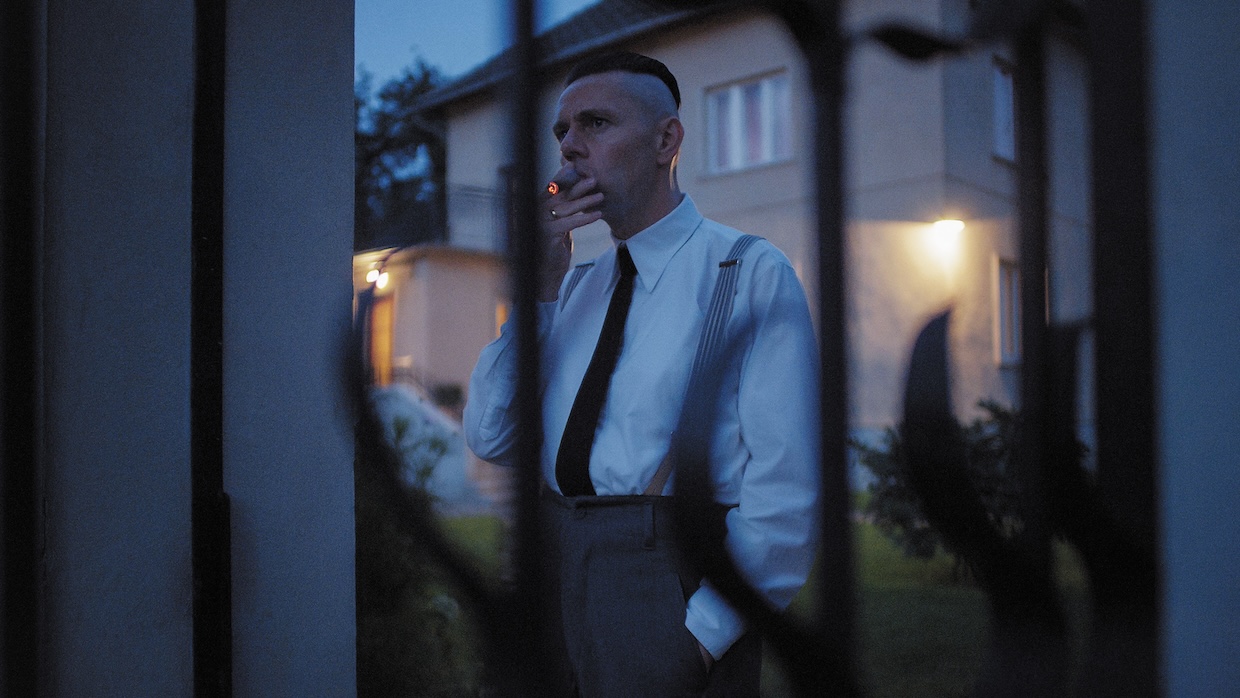 A man in pinstripe suspenders stands outside a house smoking a cigar.