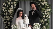 A woman in a bridal dress and a man in a tuxedo stand behind a wedding cake.