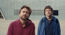 Two white men, one wearing a red hoodie and one wearing a blue zip-up, gaze upward.