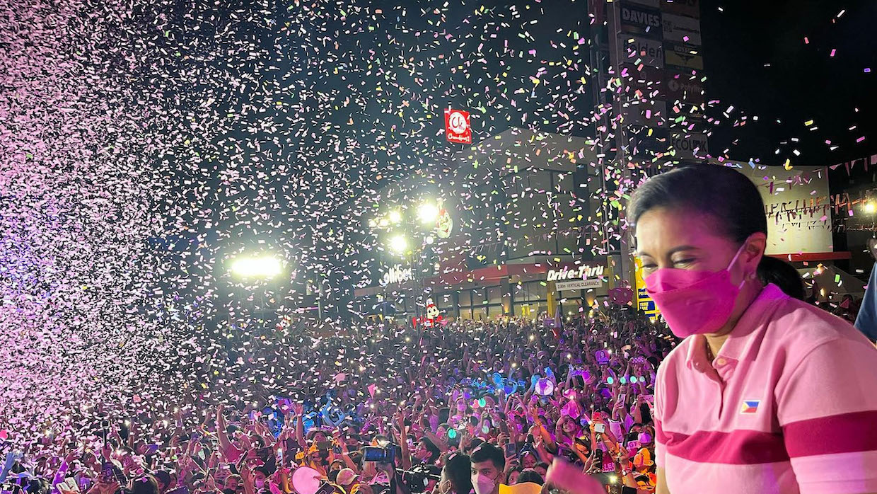 A Filipina woman wearing a pink striped polo and mask stands amid a crowd as rainbow confetti falls.