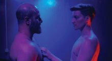 Two men, one older and balding and one younger with cropped brown hair, stand under purple and blue lights with their shirts off.