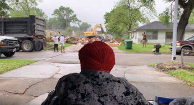An obscured person wearing a gray coat and red headwear looks across a street toward a backhoe and wooden planks.