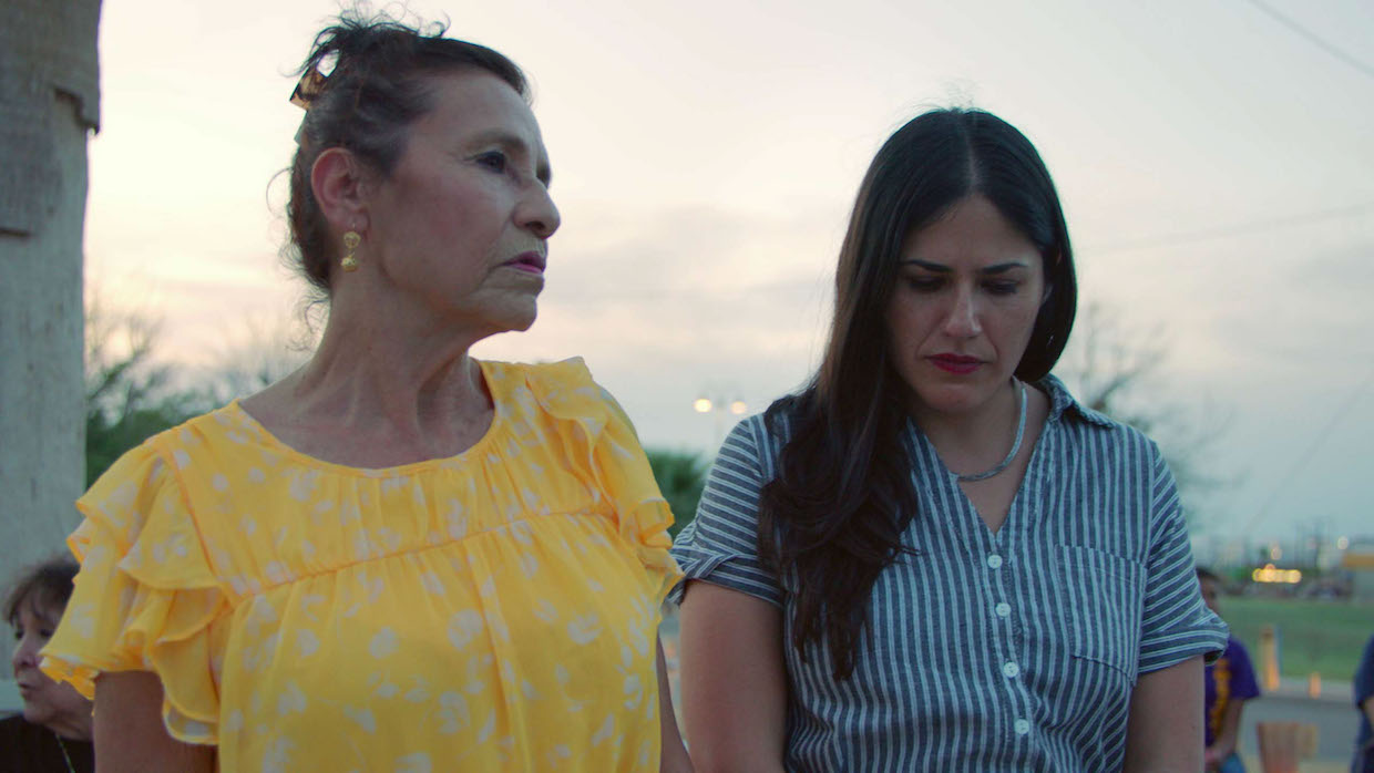 Two women stand outside during twilight. On the left, an older woman wears a ruffled yellow shirt and looks to her left. On the right, a younger woman wears a blue and white striped blouse and looks downward.