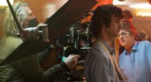 A masked cameraman adjusts focus on a crowded set, just behind a young man standing with his back to the camera.