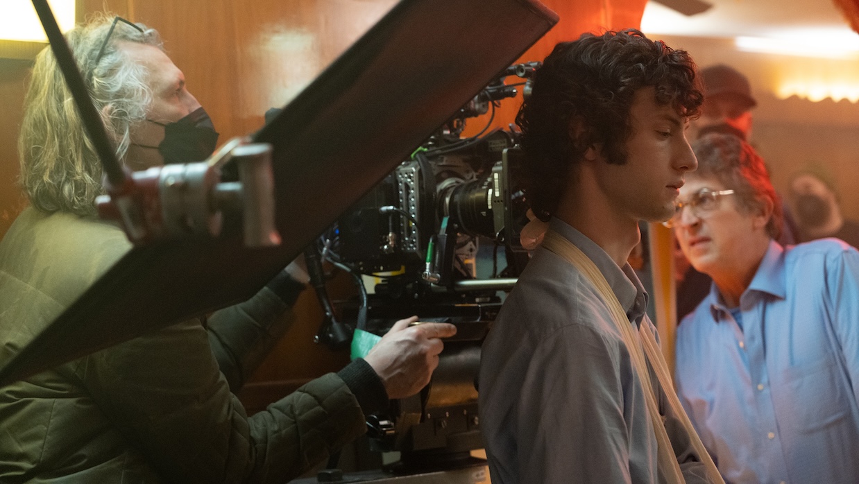 A masked cameraman adjusts focus on a crowded set, just behind a young man standing with his back to the camera.