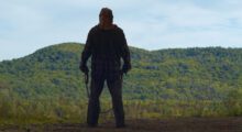 A masked man armed with an obscured weapon stands against a forested backdrop.