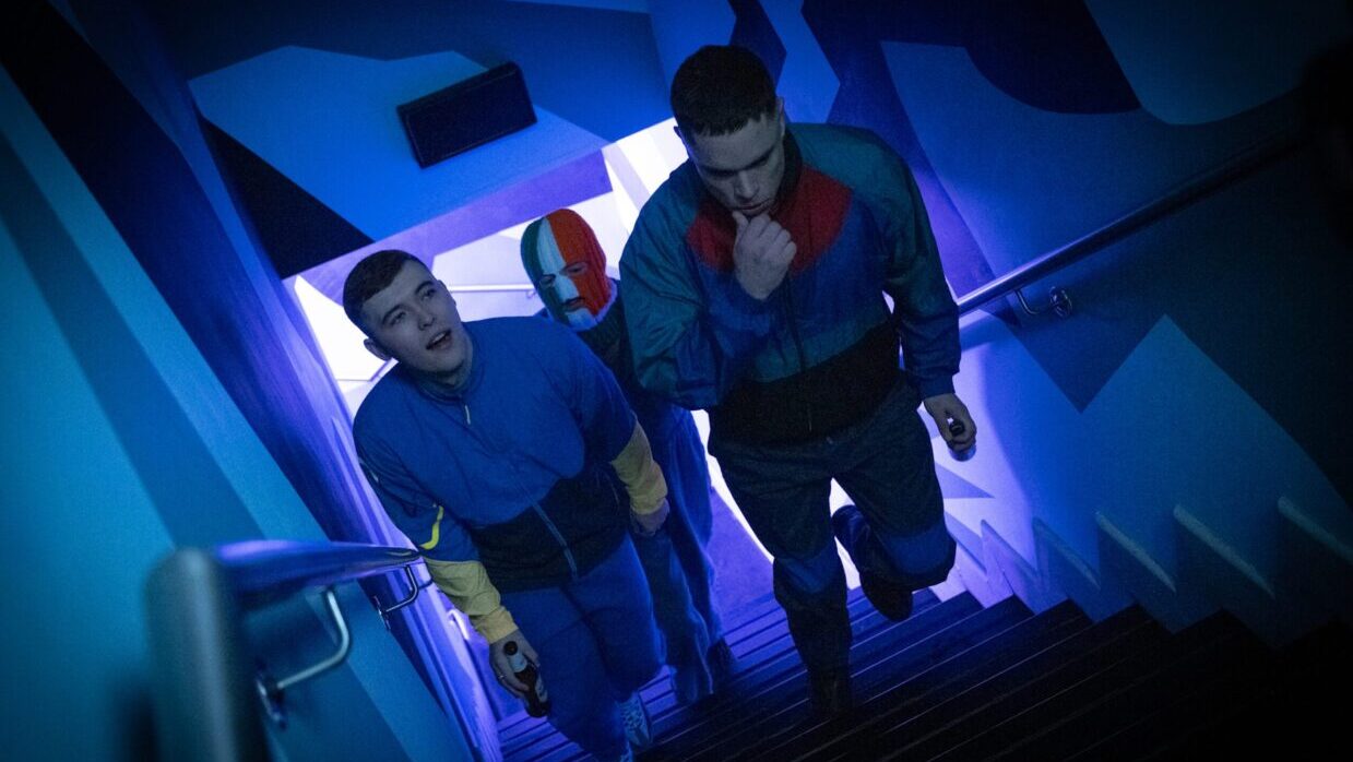 Three Irish men dressed in blue ascend a staircase. The man in the back is wearing a red, white and blue ski mask.