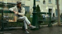 A man with a paper takeout bag from McDonald's sits on a park bench in daylight.