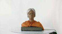 BINA48, a humanoid AI consisting of shoulders and head with dark skin and gray hair, is sitting on a table.