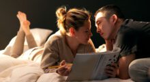On a white bedspread, a white woman holding a magazine and an Asian man look into one another's eyes.