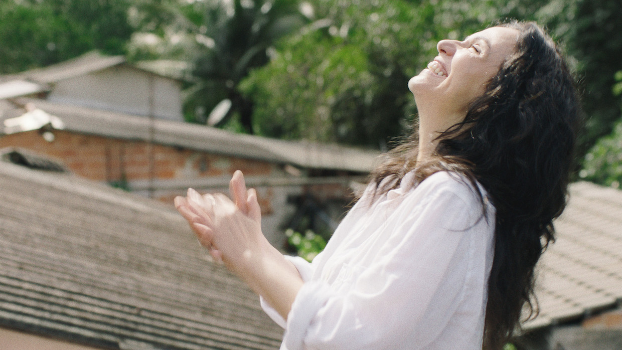 A white woman with long, curly dark hair wears a white tunic and stands smiling in the sunlight.