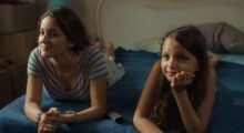 Two Latina girls lying on a blue bedspread are propping themselves up with their arms.